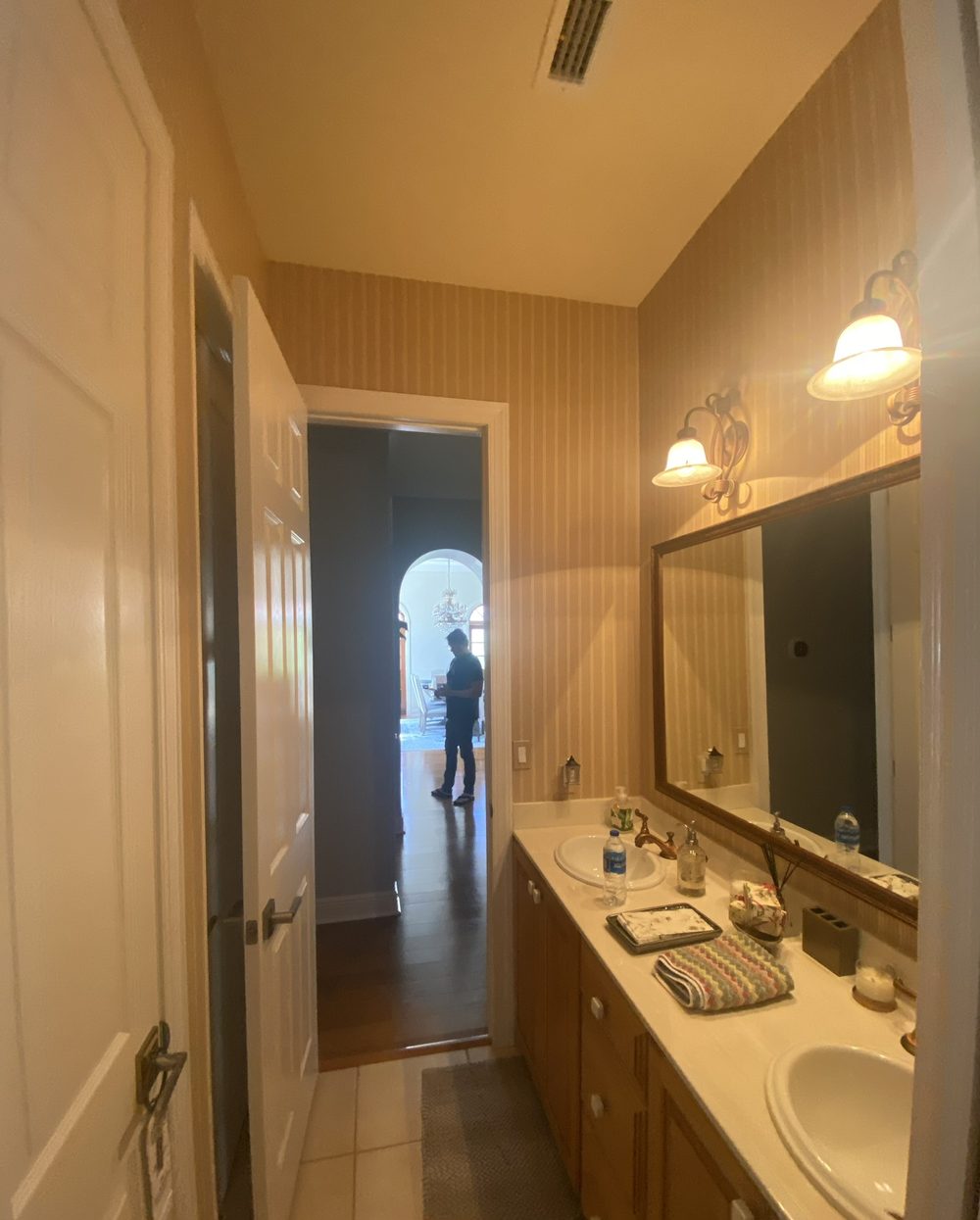 Guest bathroom before remodel — dated double-sink vanity with striped wallpaper and brass mirror frame